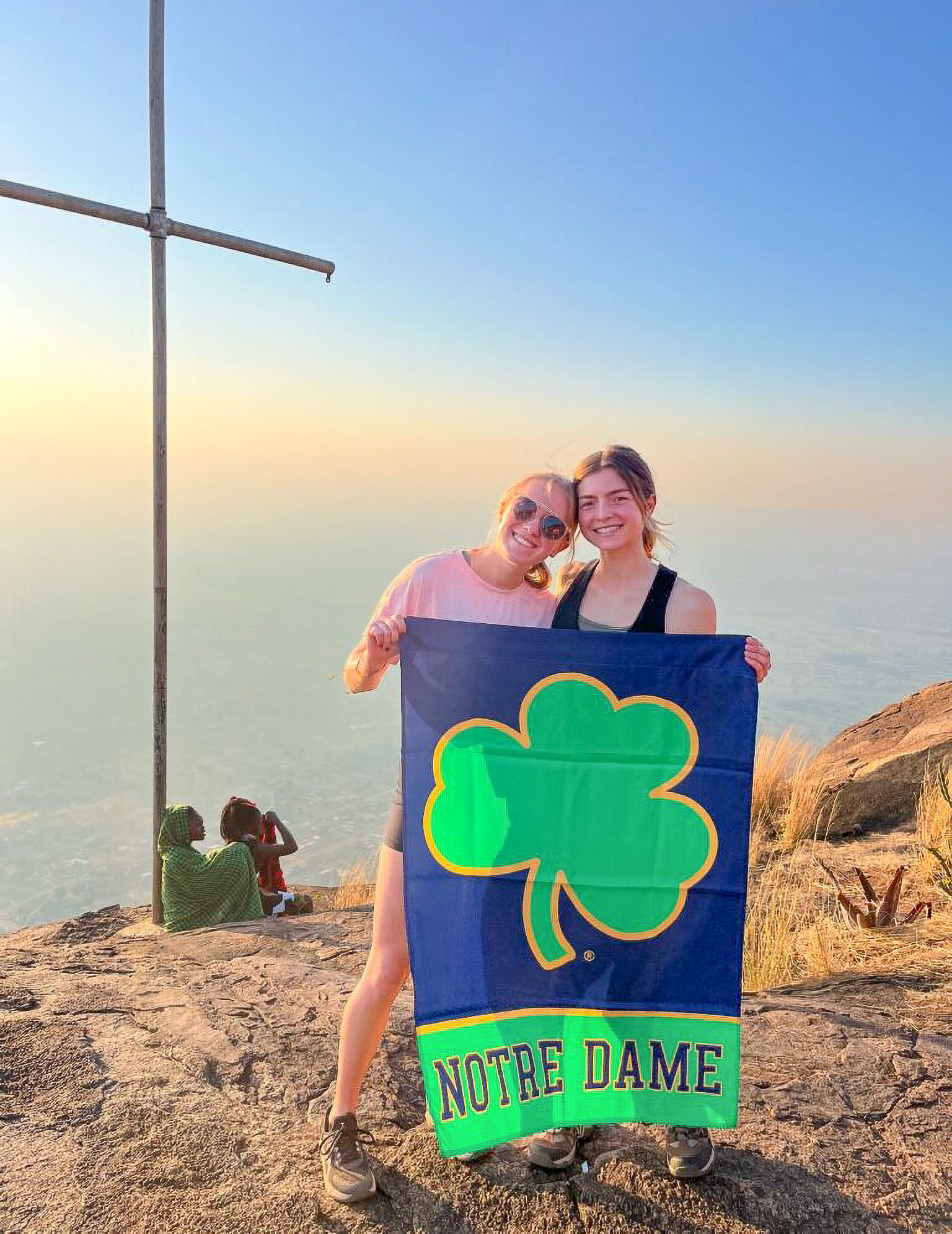 Two people holding a Notre Dame flag at a rocky summit, with a large cross and distant fog under a clear sky.