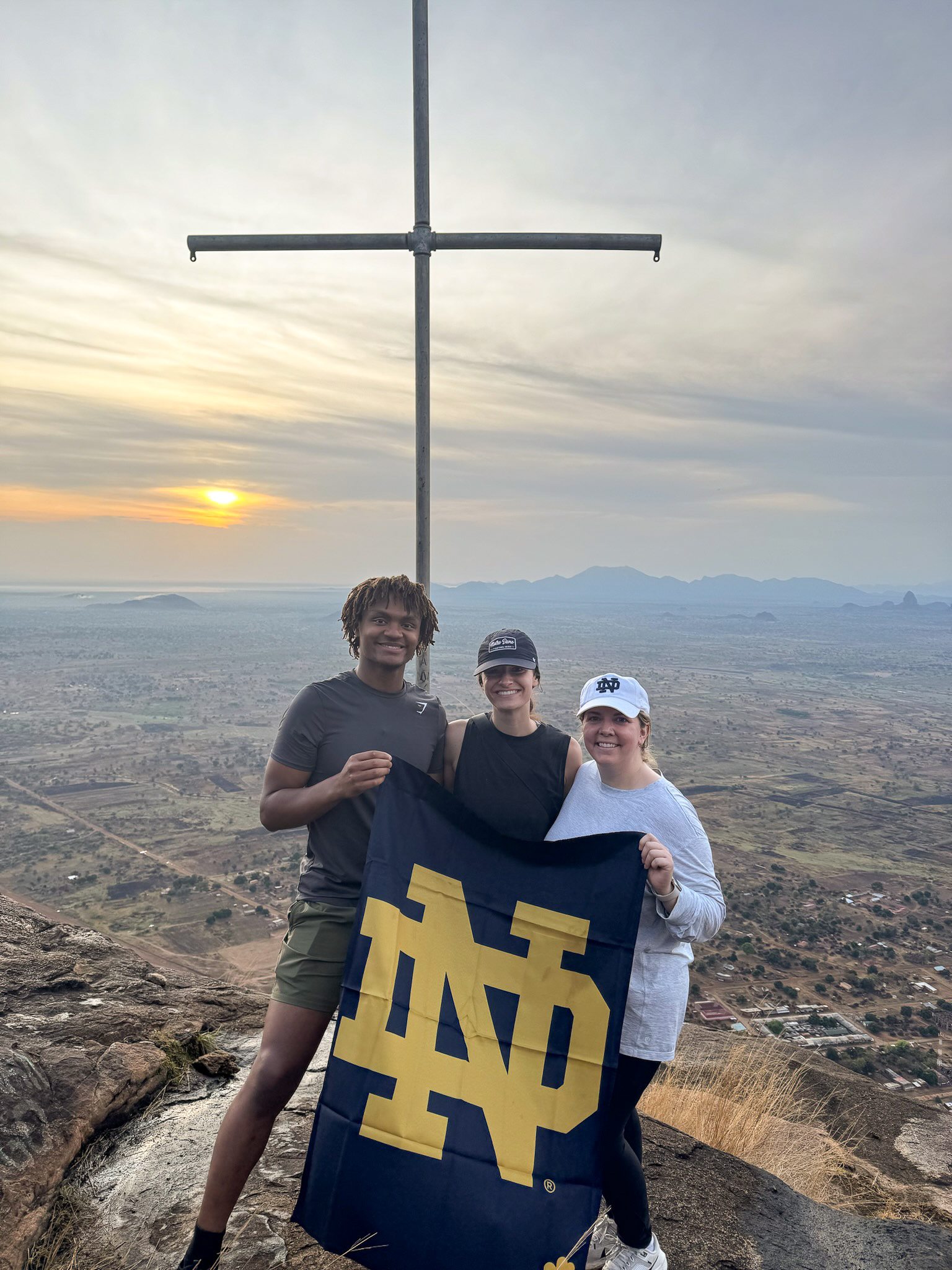 Three people hold a Notre Dame flag at a scenic hilltop with a cross, overlooking a vast landscape at sunset.