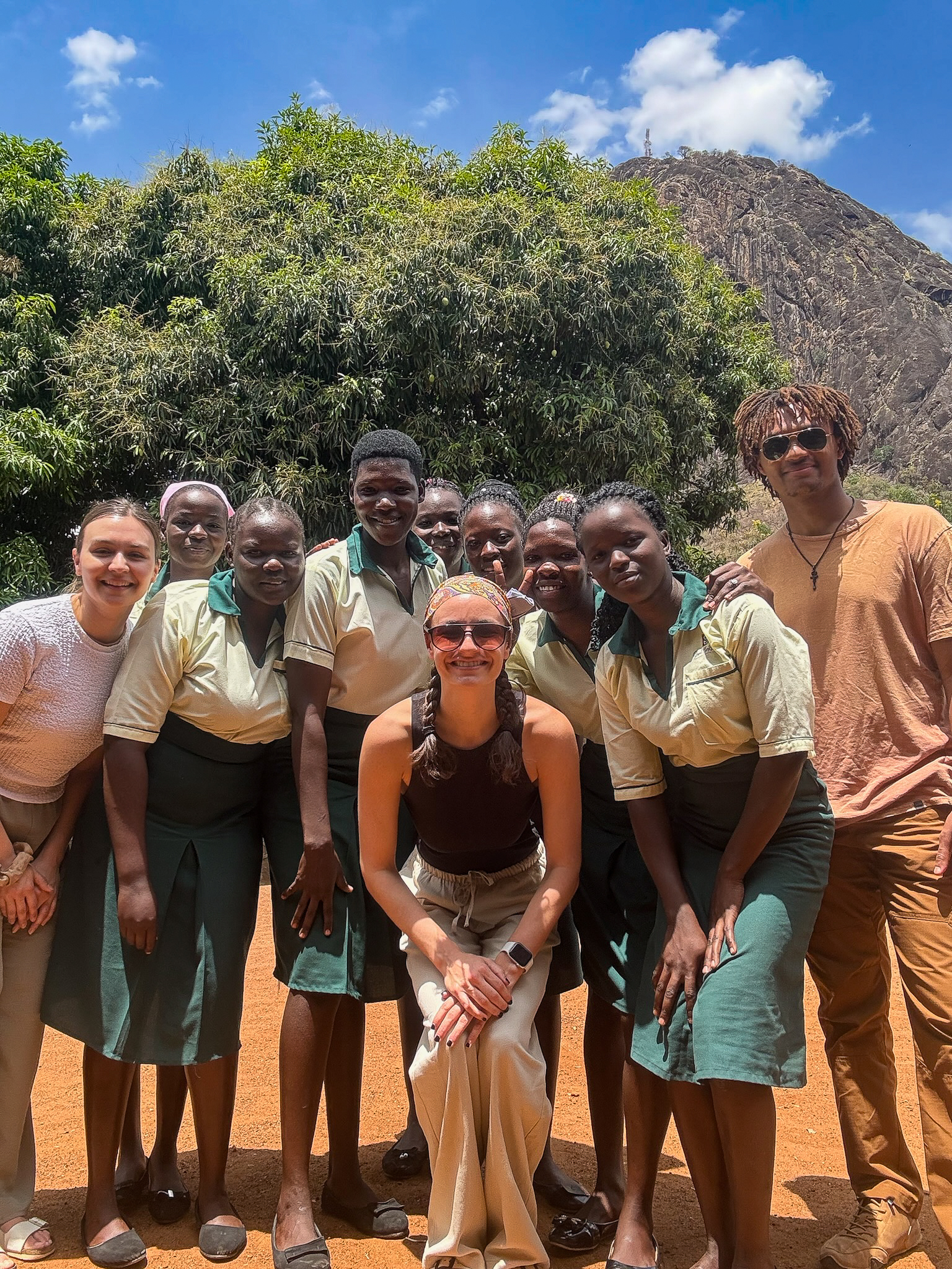 A group of people stands together outdoors in front of a large tree and rock formation on a sunny day, all smiling at the camera.