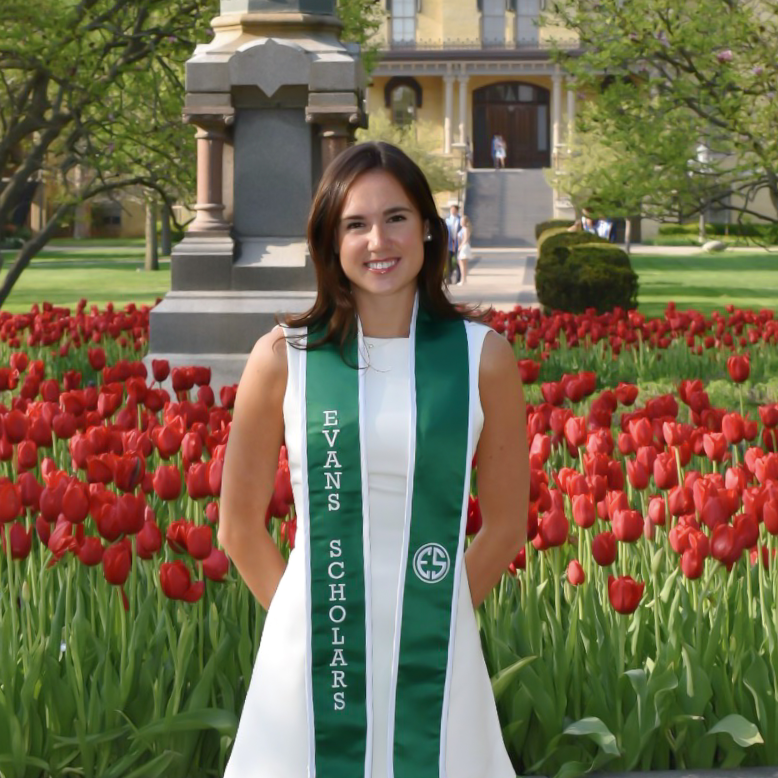 A woman in a white dress and green Evans Scholars stole stands in front of red tulips, with a stone monument and building in the background.