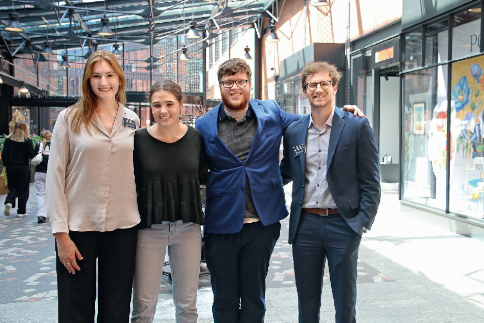 Four people stand together smiling in an outdoor walkway with glass roof, wearing business casual attire.