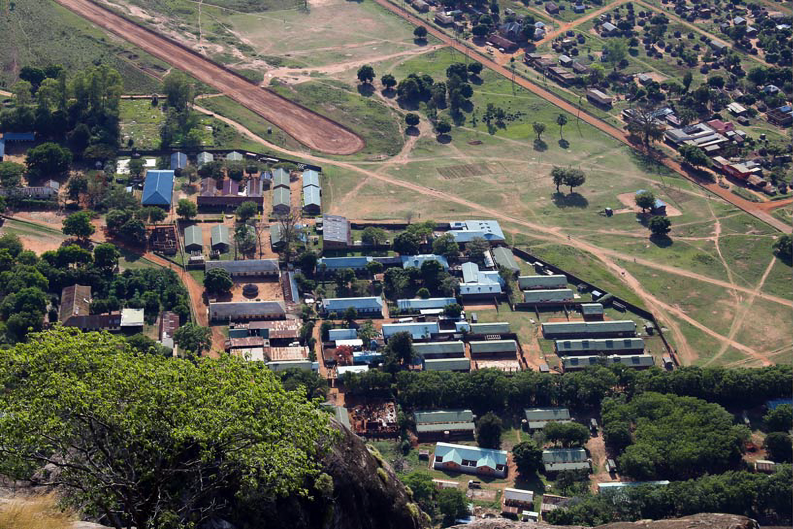 Aerial view of a small town with clustered buildings, roads, and surrounding greenery, set against a backdrop of open fields and hills.