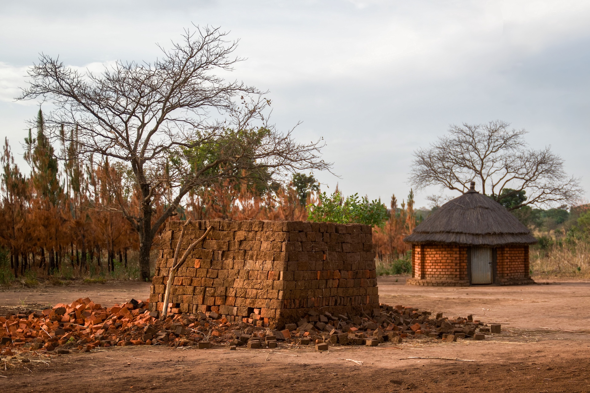 A brick kiln with scattered bricks in the foreground, and a round, thatched-roof hut in the background, set in a rural landscape.