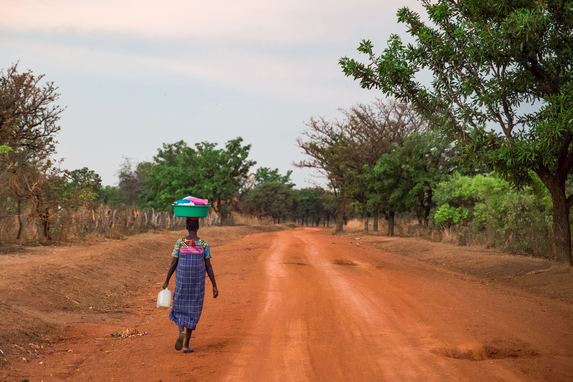 A person walks down a rural dirt road carrying a container on their head and a jug in hand, surrounded by trees and a clear sky.