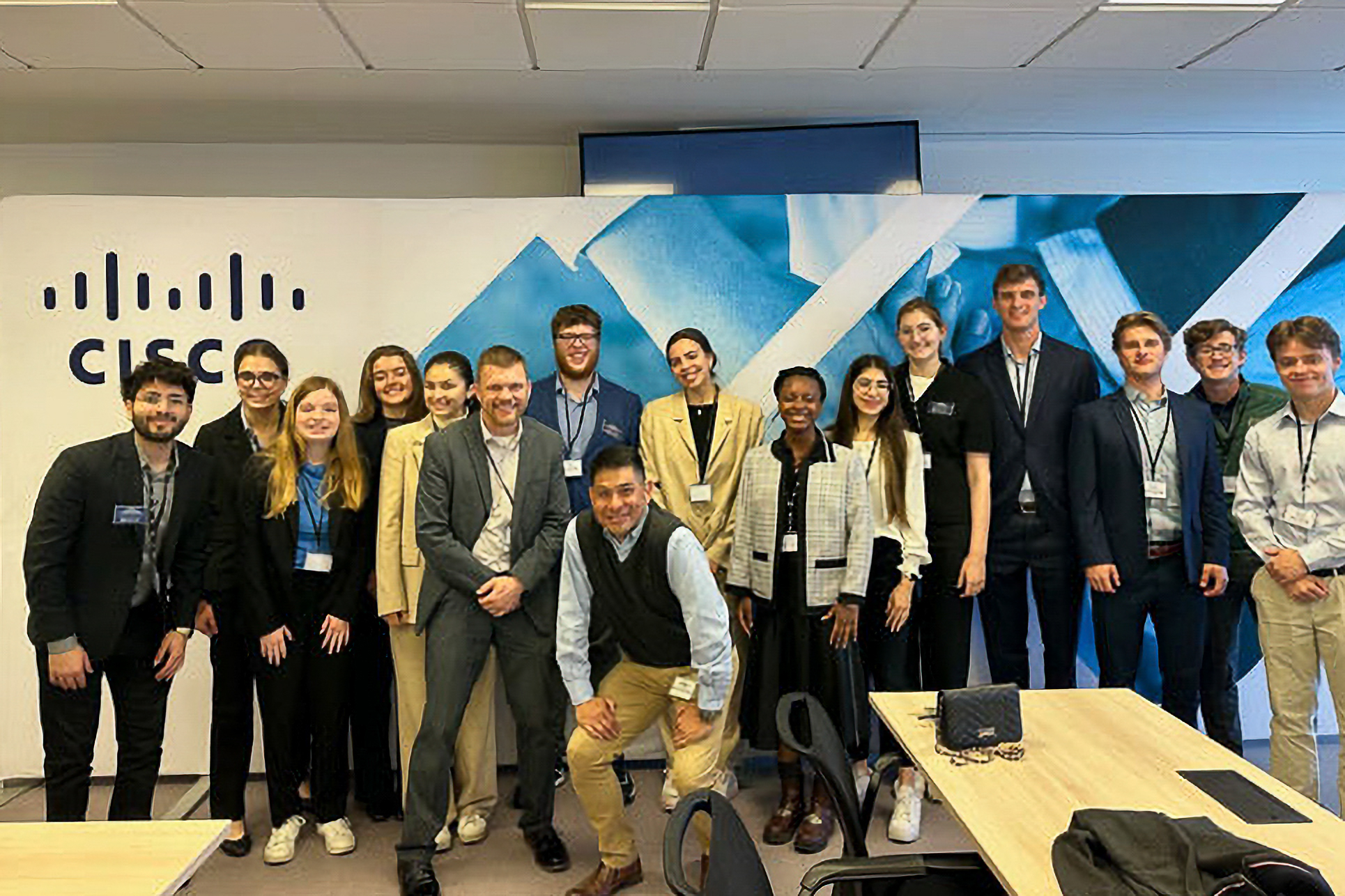 A diverse group of people posing and smiling in front of a Cisco sign in a conference room, with tables and chairs in the foreground.
