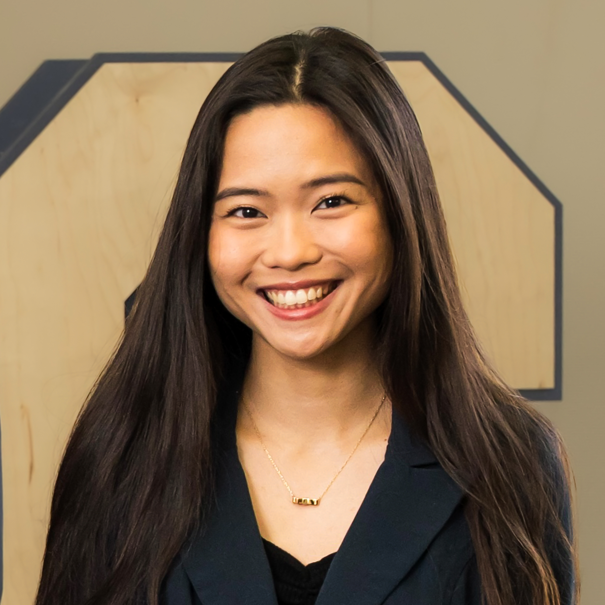 Smiling woman with long dark hair, wearing a black blazer and necklace, in front of a geometric wooden backdrop.
