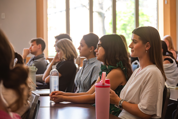 A group of people attentively listening in a well-lit room, with water bottles and notes on the table in front of them.