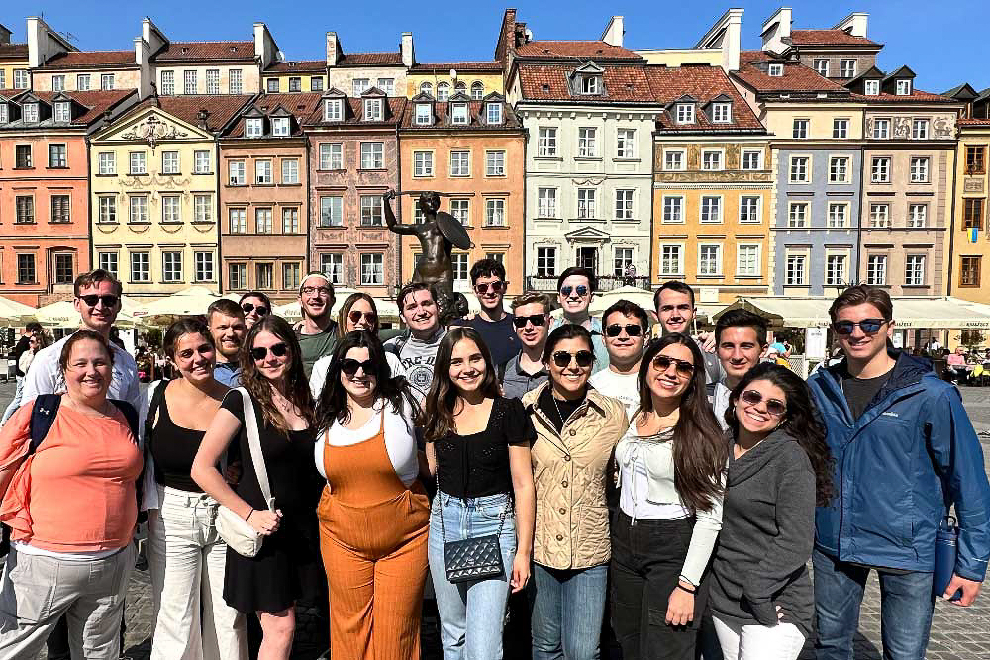 A group of smiling people poses in front of colorful, historic buildings under a clear blue sky.