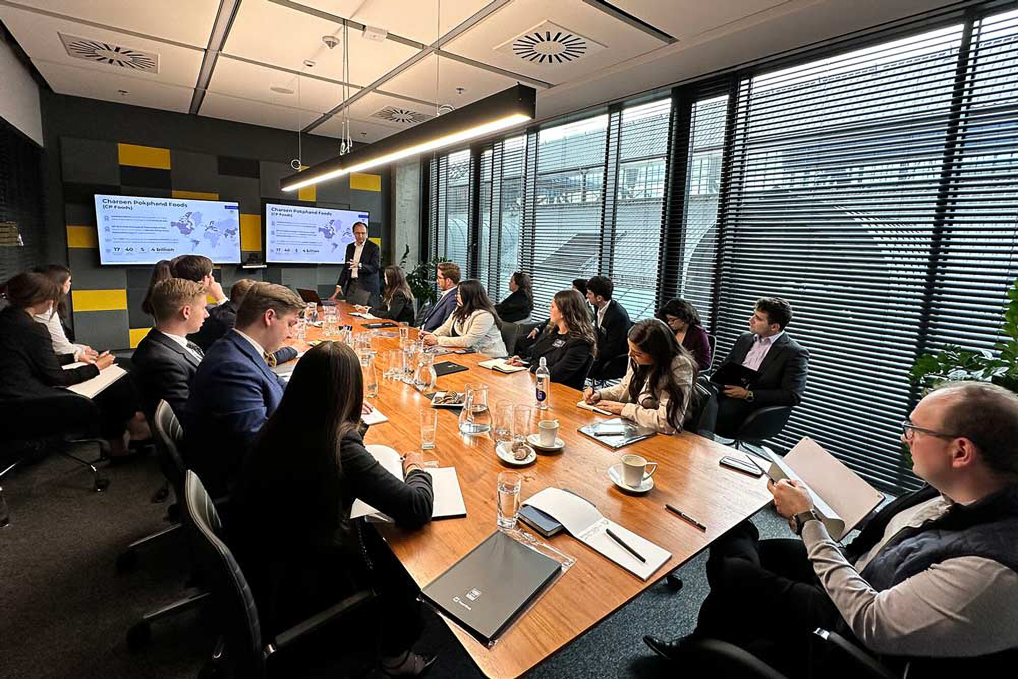 People seated around a conference table in a modern office, watching a presentation on two screens. The room has large windows with blinds.