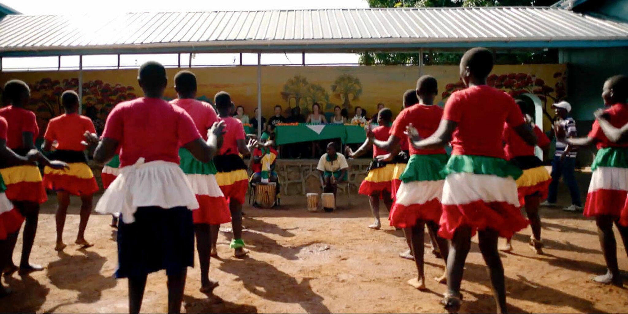 A group of dancers in red tops and colorful skirts perform outdoors in front of an audience seated under a shelter.