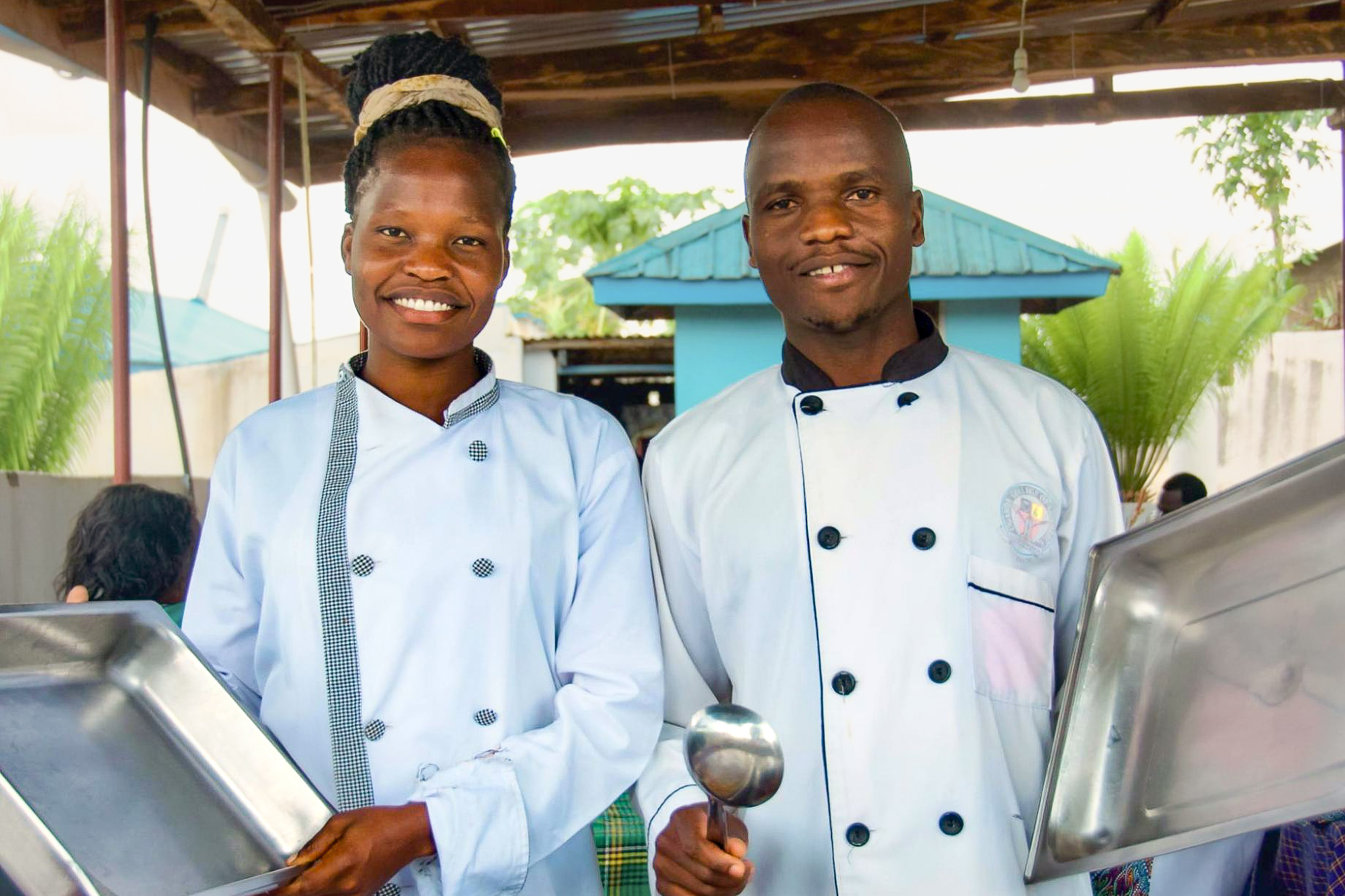 Two chefs in white uniforms, smiling and holding cooking trays and a ladle, stand under a shelter with greenery in the background.