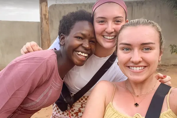 Three smiling friends take a selfie outdoors, two embracing the third, with a wall and a tree in the background.