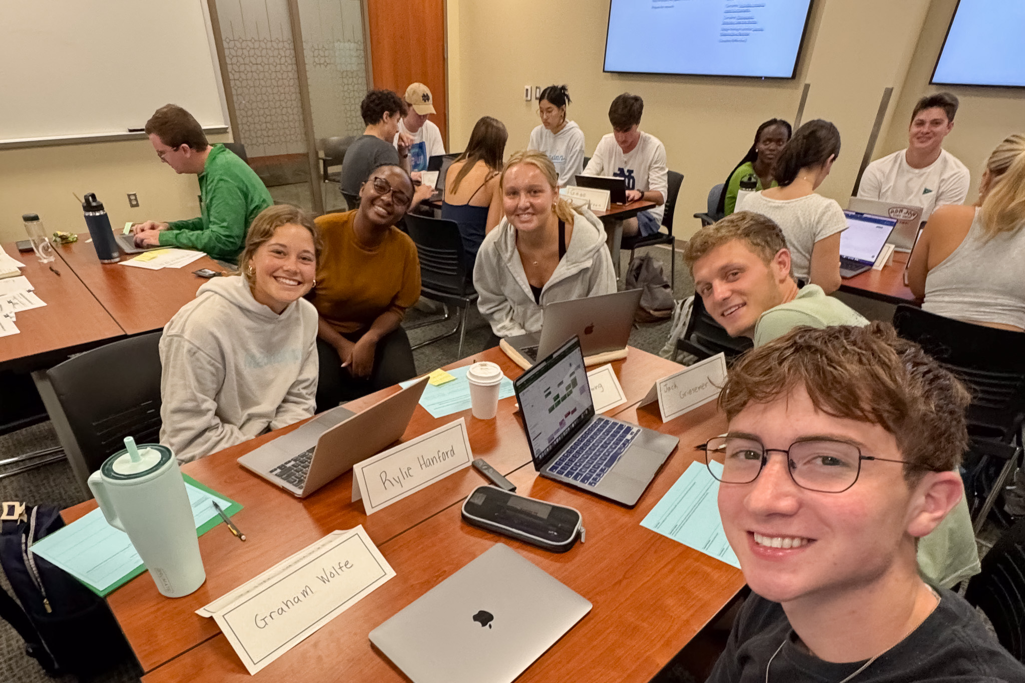 A group of students smiling and working on laptops in a classroom setting, seated around wooden desks with nameplates visible.