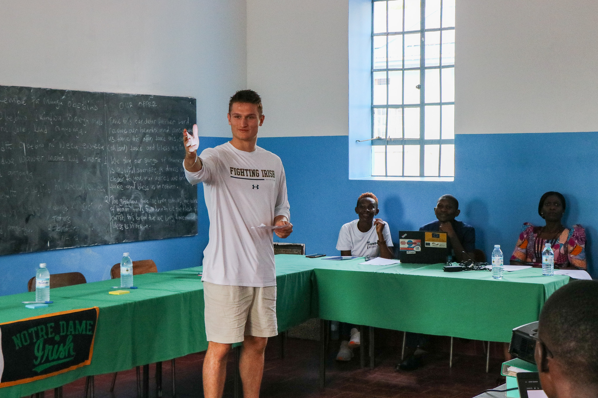 A person in a "Fighting Irish" shirt speaks to a group in a classroom with a chalkboard and green tables.