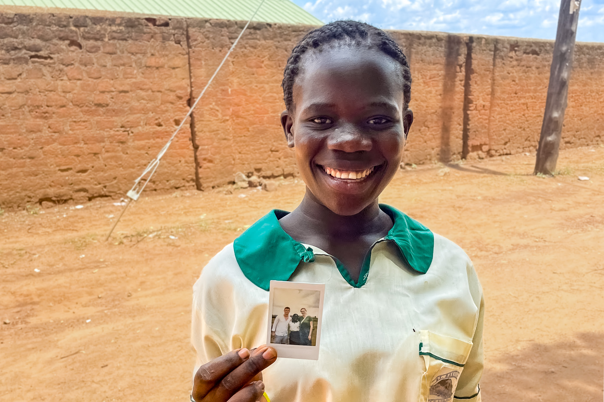 Smiling girl in a school uniform holds a small photo, standing outside with a brick wall and clear sky in the background.
