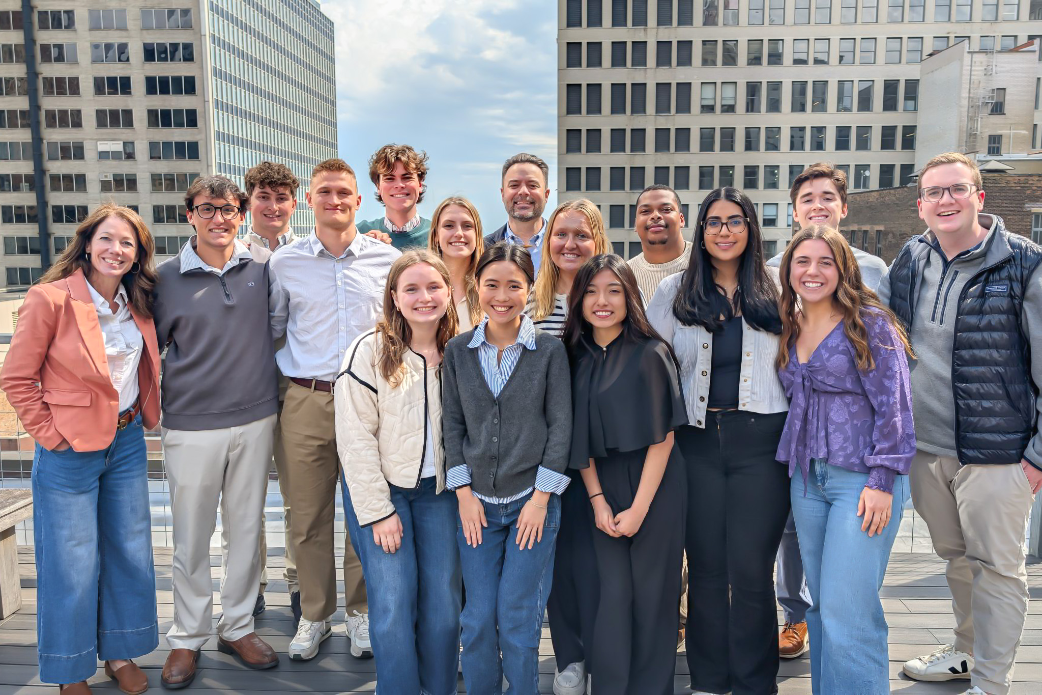 A group of people smiling on a rooftop with city buildings in the background. They're casually dressed, standing closely together.