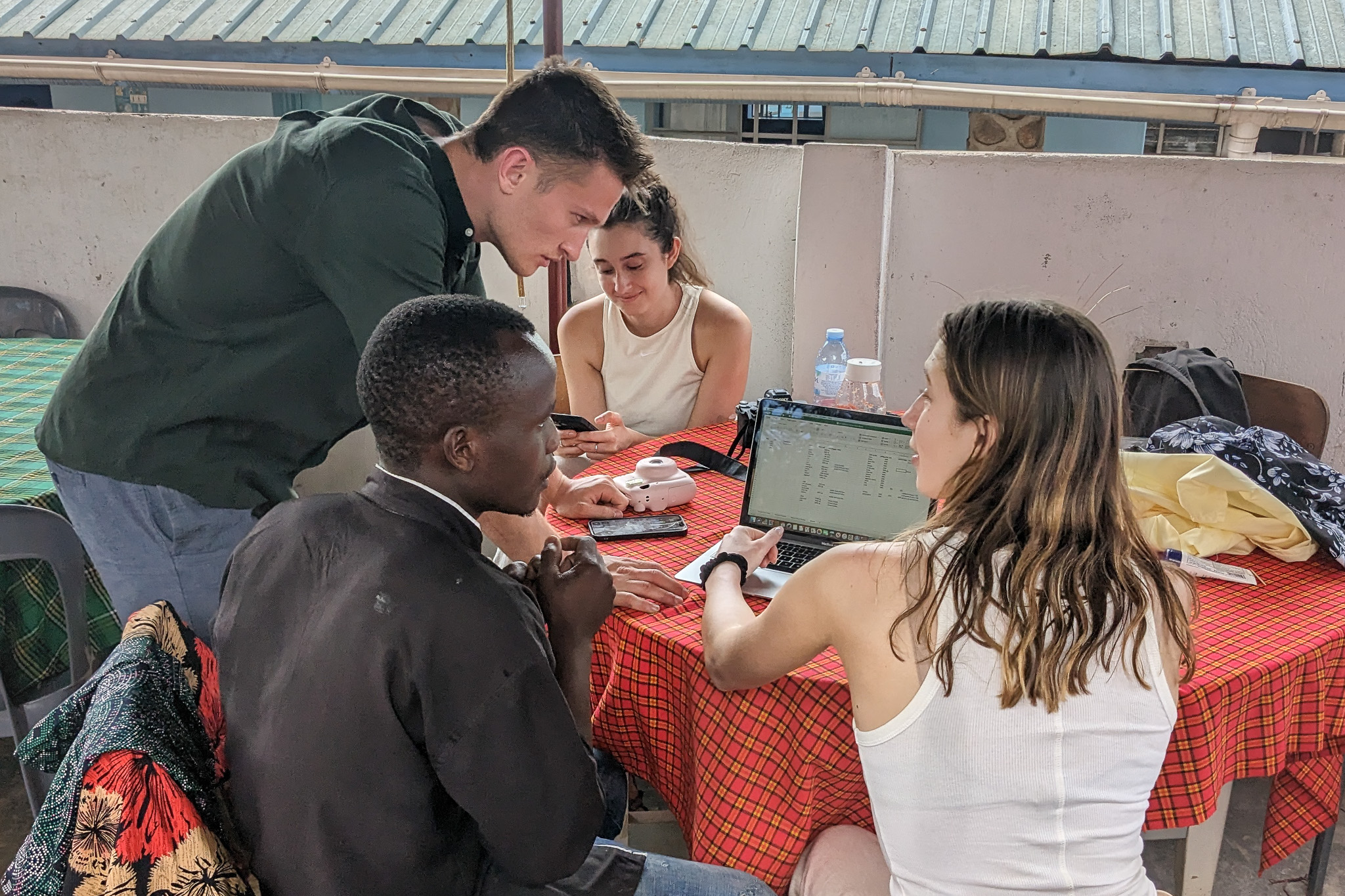 Four people collaborate around a table with a laptop, documents, and phones, under an open shed with a corrugated metal roof.
