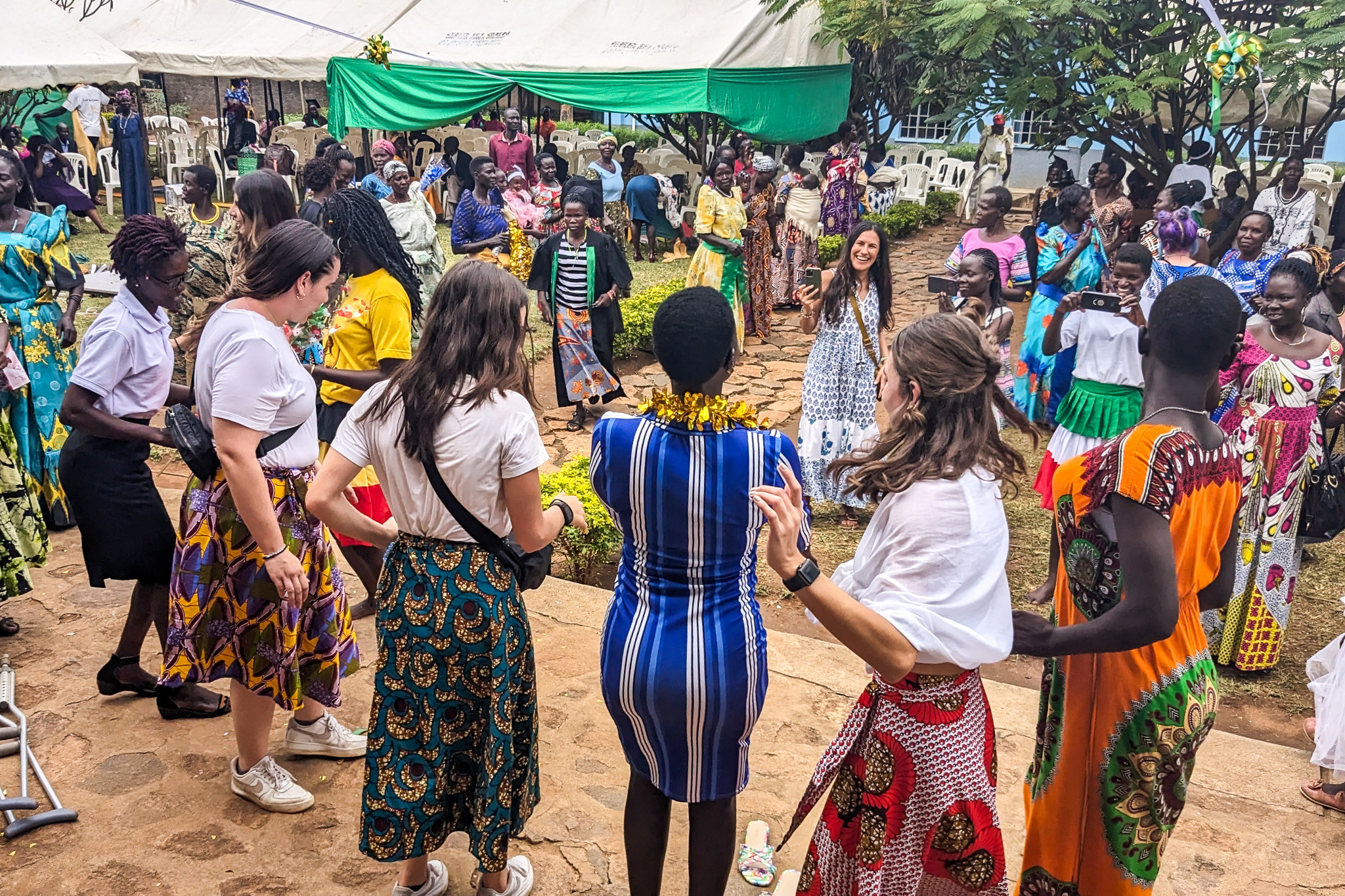 A diverse group of people joyfully dancing together outdoors, surrounded by tents and onlookers capturing the moment on their phones.