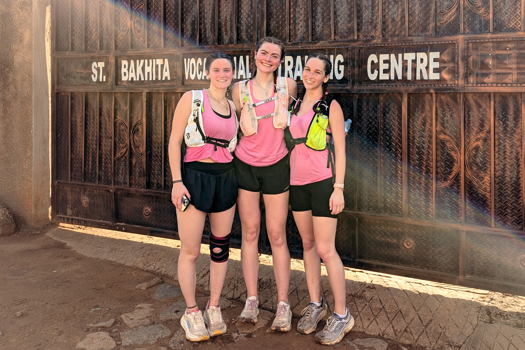 Three women in matching pink tops and shorts stand smiling in front of a gate labeled "St. Bakhita Vocational Training Centre."