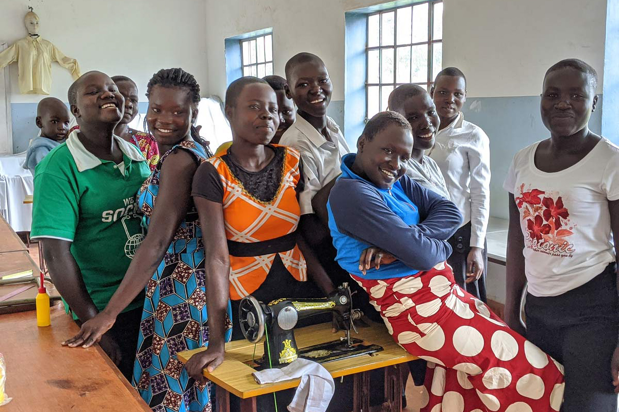 A group of smiling people gathered around a sewing machine in a bright room with barred windows.