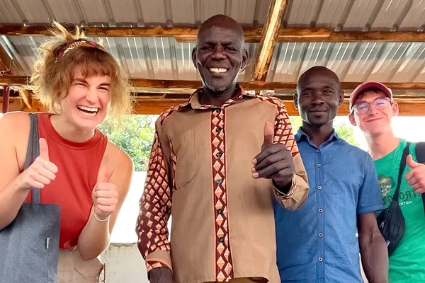 Four people smiling and giving thumbs up, standing under a wooden and metal roof.