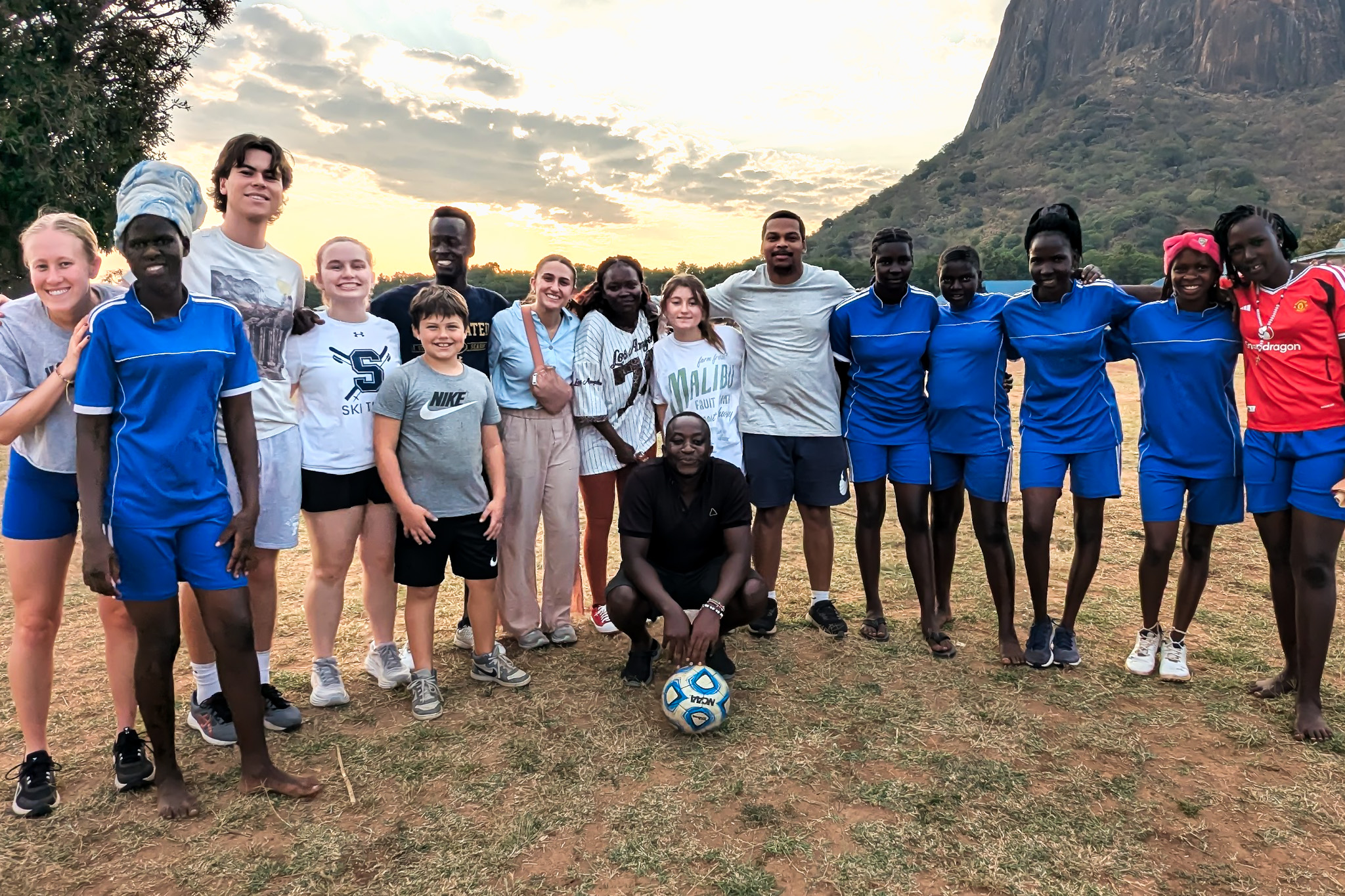 A group of people in casual and sportswear pose outdoors on a grassy field with a mountain and sunset in the background.