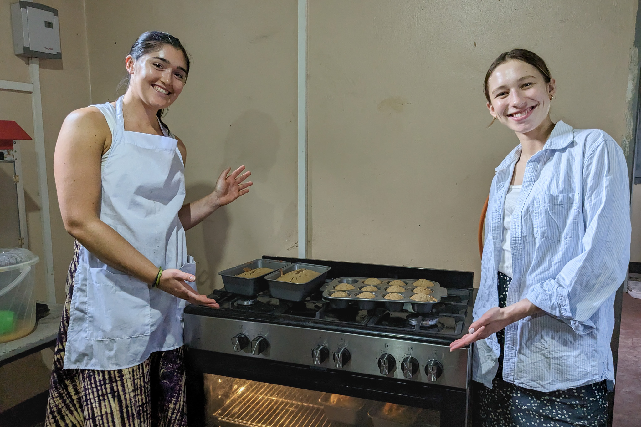 Two women smiling and pointing to freshly baked bread and muffins on a stove in a kitchen.