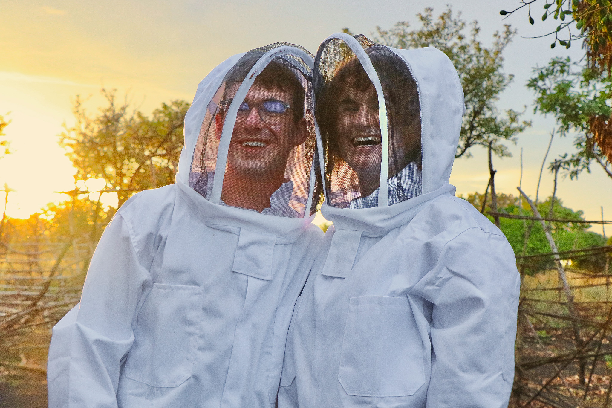 Two people in white beekeeping suits and veils smile at sunset, with trees and a wooden fence in the background.