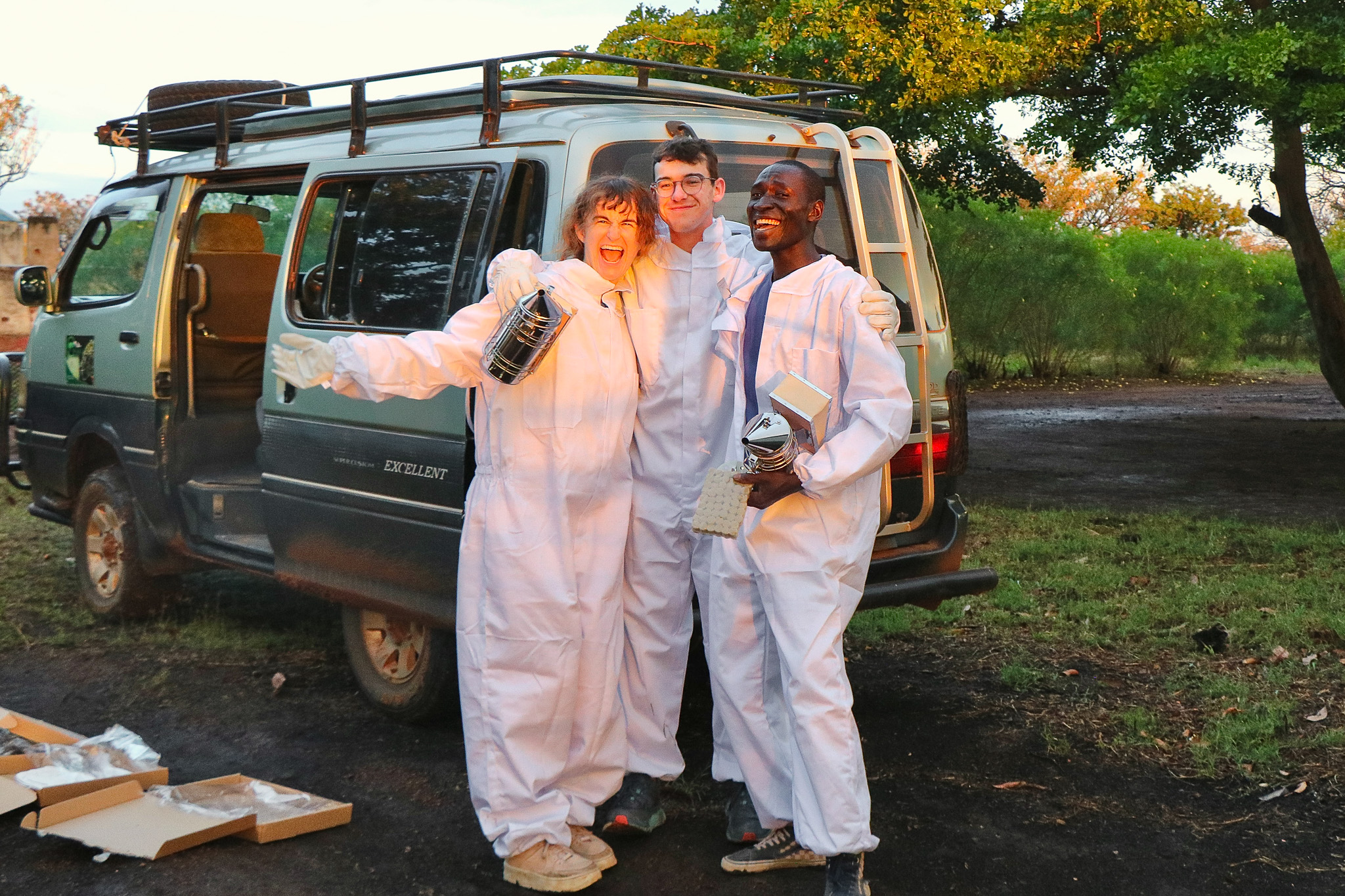 Three people in white protective suits stand smiling in front of a van, surrounded by greenery and boxes on the ground.