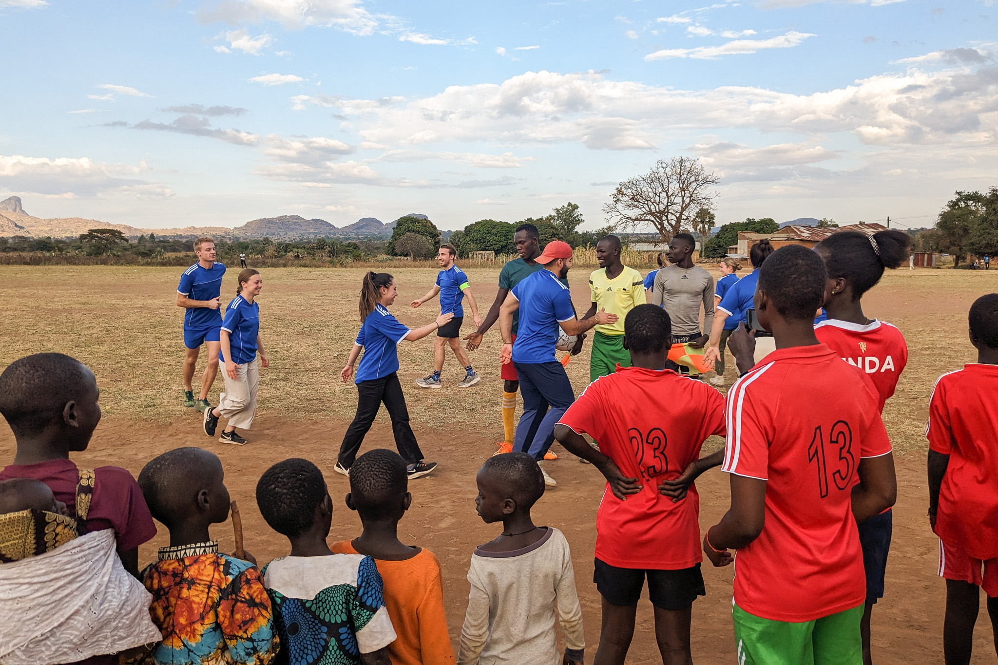 People in blue shirts greet others on a field, while children in colorful clothes watch. Mountains and clouds are visible in the background.