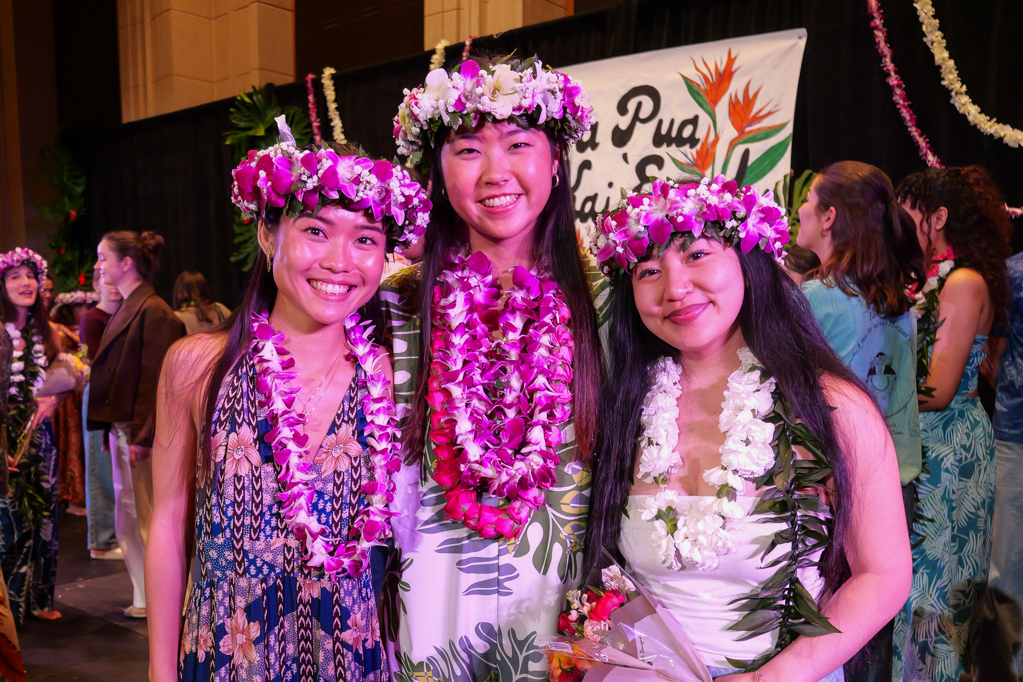 Three women wearing floral leis and headdresses smile at a Na Pua Kai 'Ewalu event, standing in front of a decorative sign.