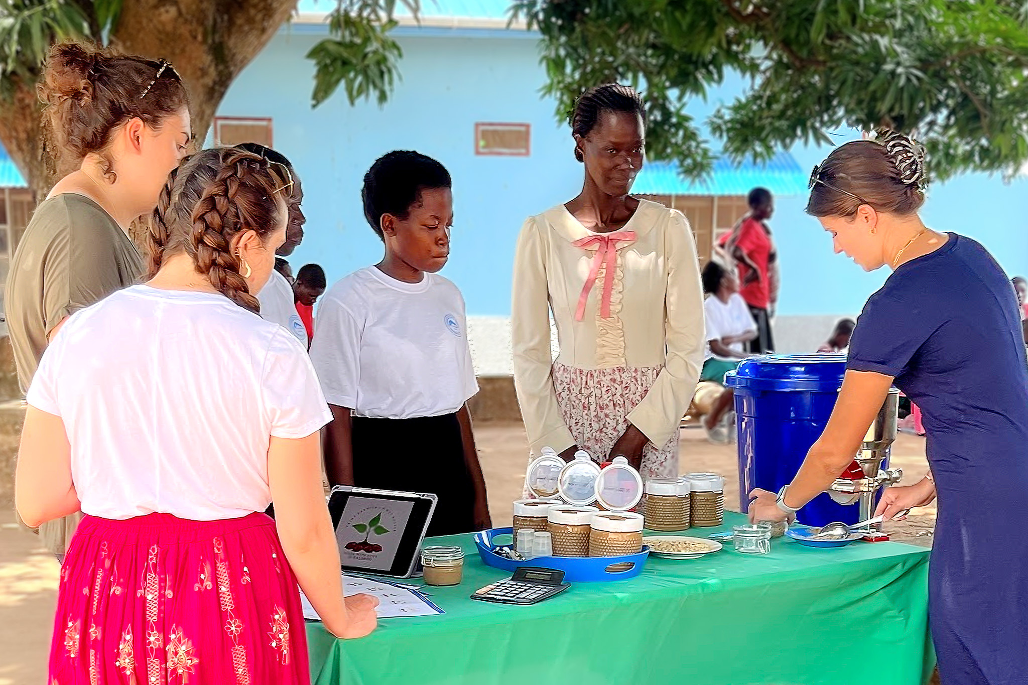 A group of people stand around a table with jars and a blue container, under a tree in an outdoor setting.