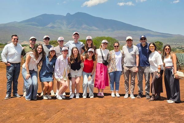 A group of people pose for a photo outdoors, wearing casual clothing and hats, with a mountain visible in the background under a clear blue sky.