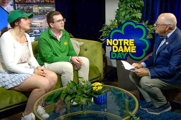 Three people are seated in a studio setting with "Notre Dame Day" signage. Two are on a couch, and one is interviewing them.