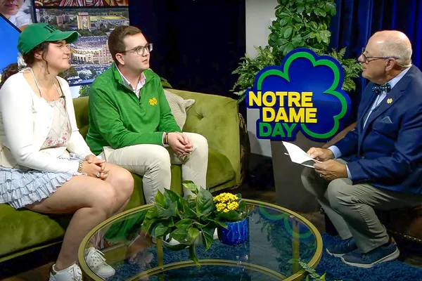 Three people are seated in a studio setting with "Notre Dame Day" signage. Two are on a couch, and one is interviewing them.