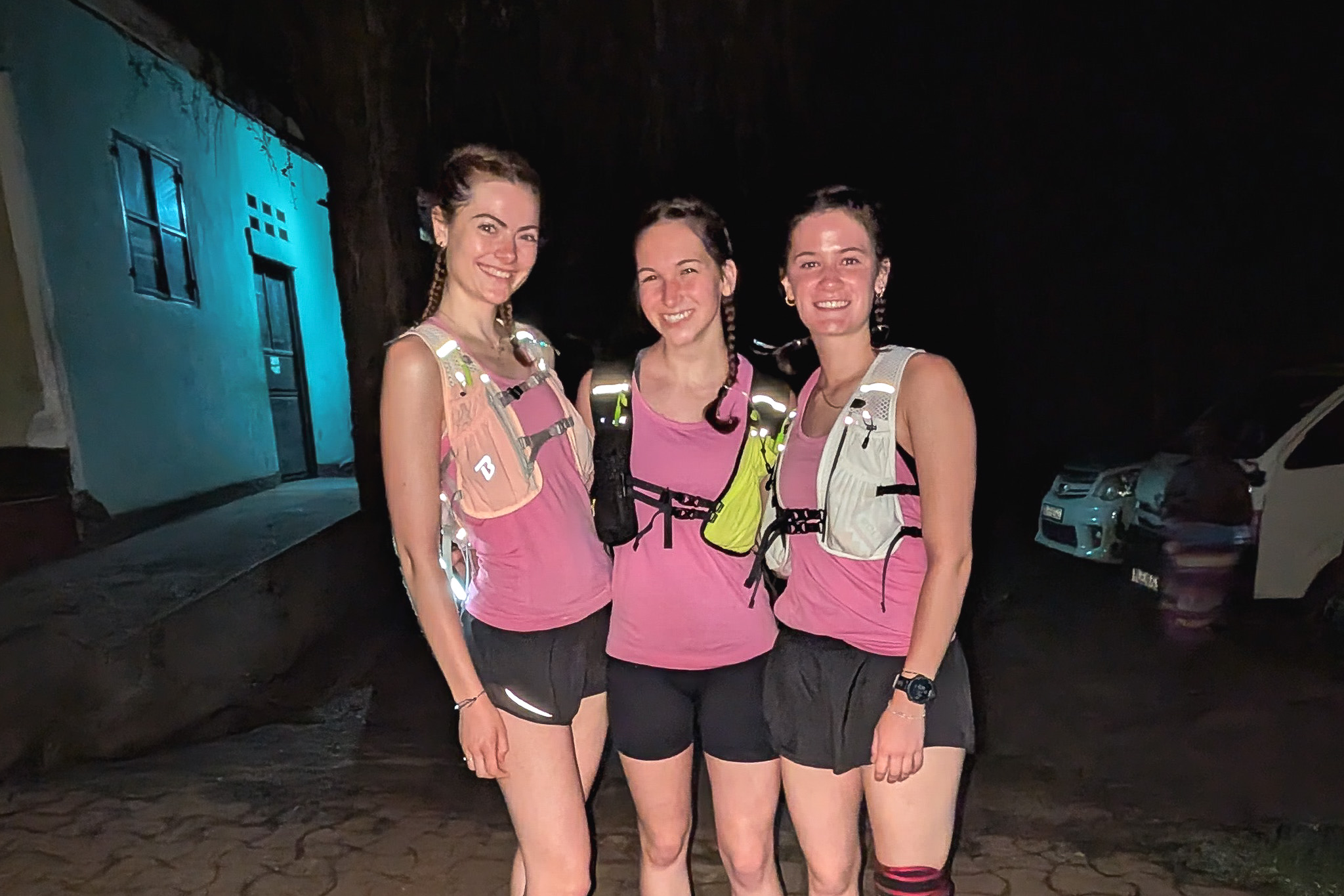 Three smiling women in matching pink tops and shorts pose at night with running gear, in front of a building and parked cars.