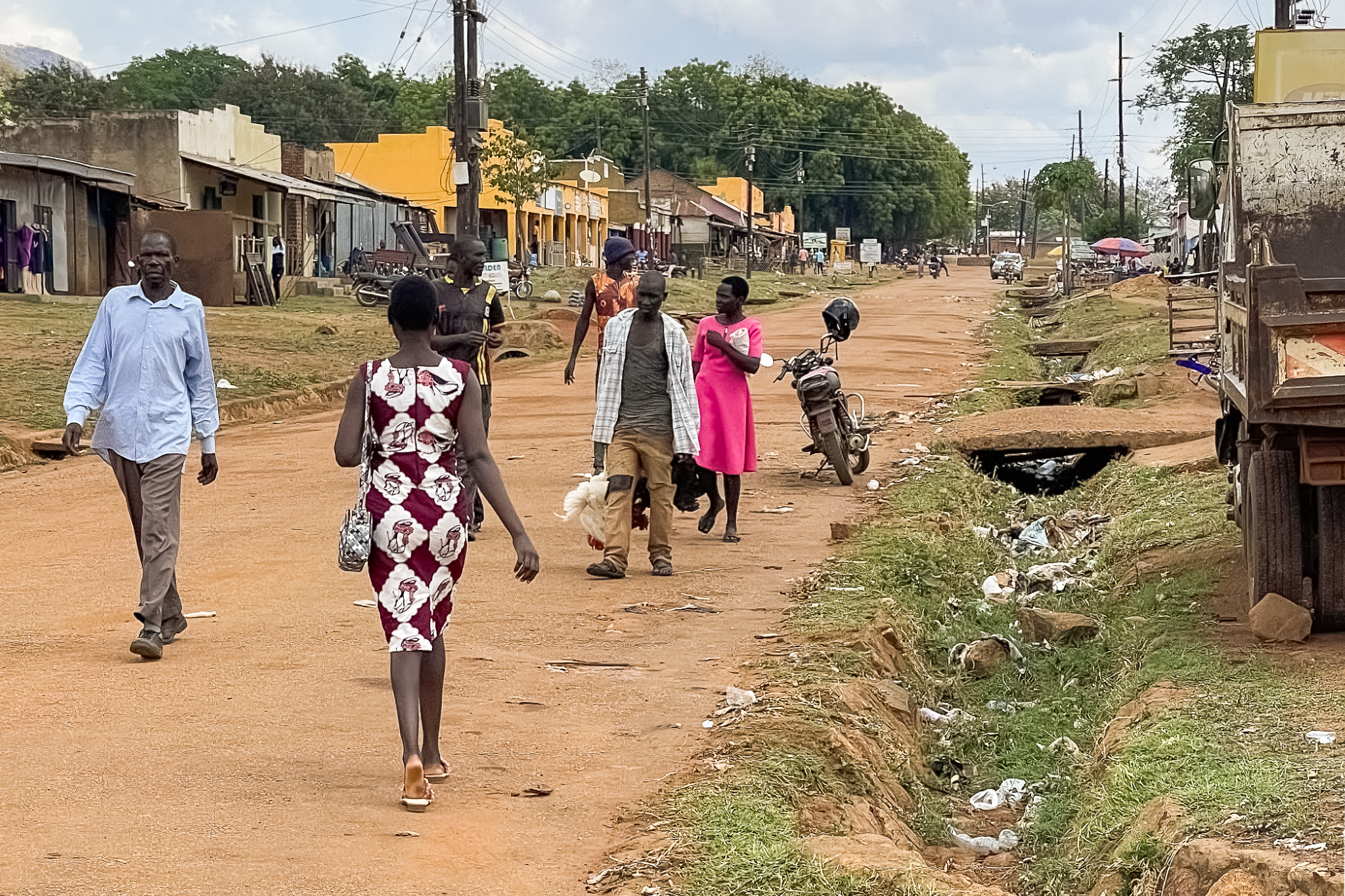 People walking on a dirt road in a rural area with shops and buildings on the side. A motorcycle is parked nearby.