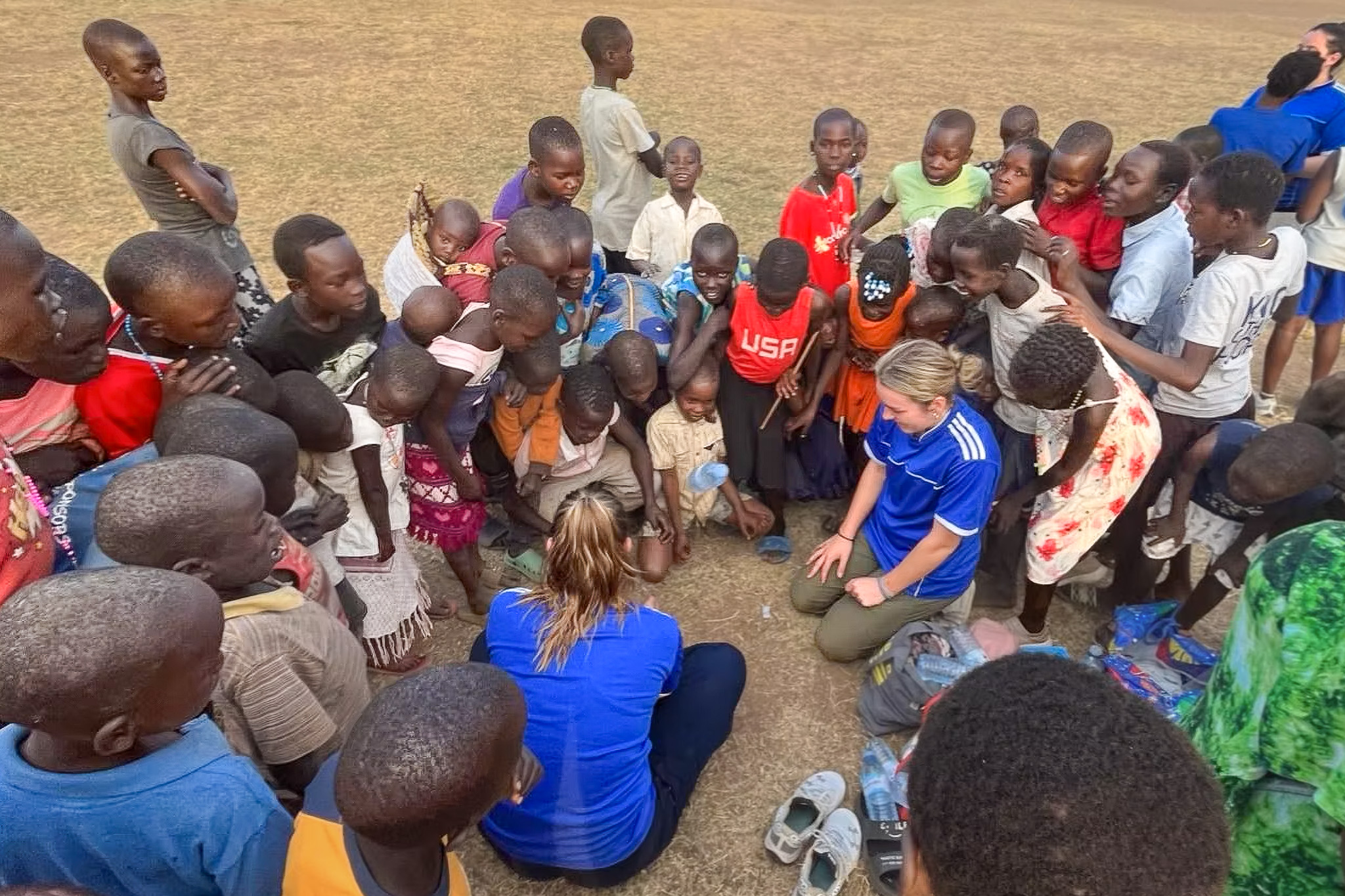 A group of children gathered around two people in blue shirts sitting on the ground, interacting and engaging with the kids.