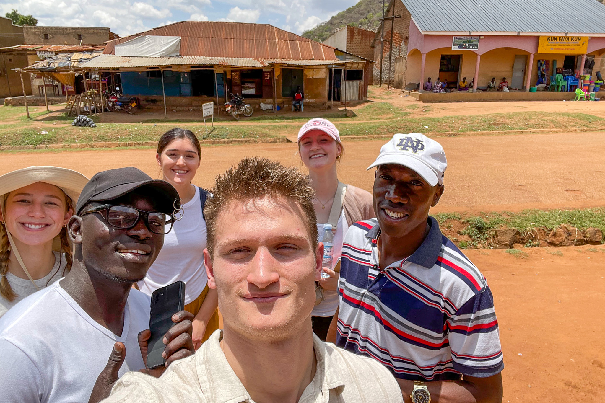 A group of six people smiling for a selfie in a rural area with buildings and a dirt road in the background.