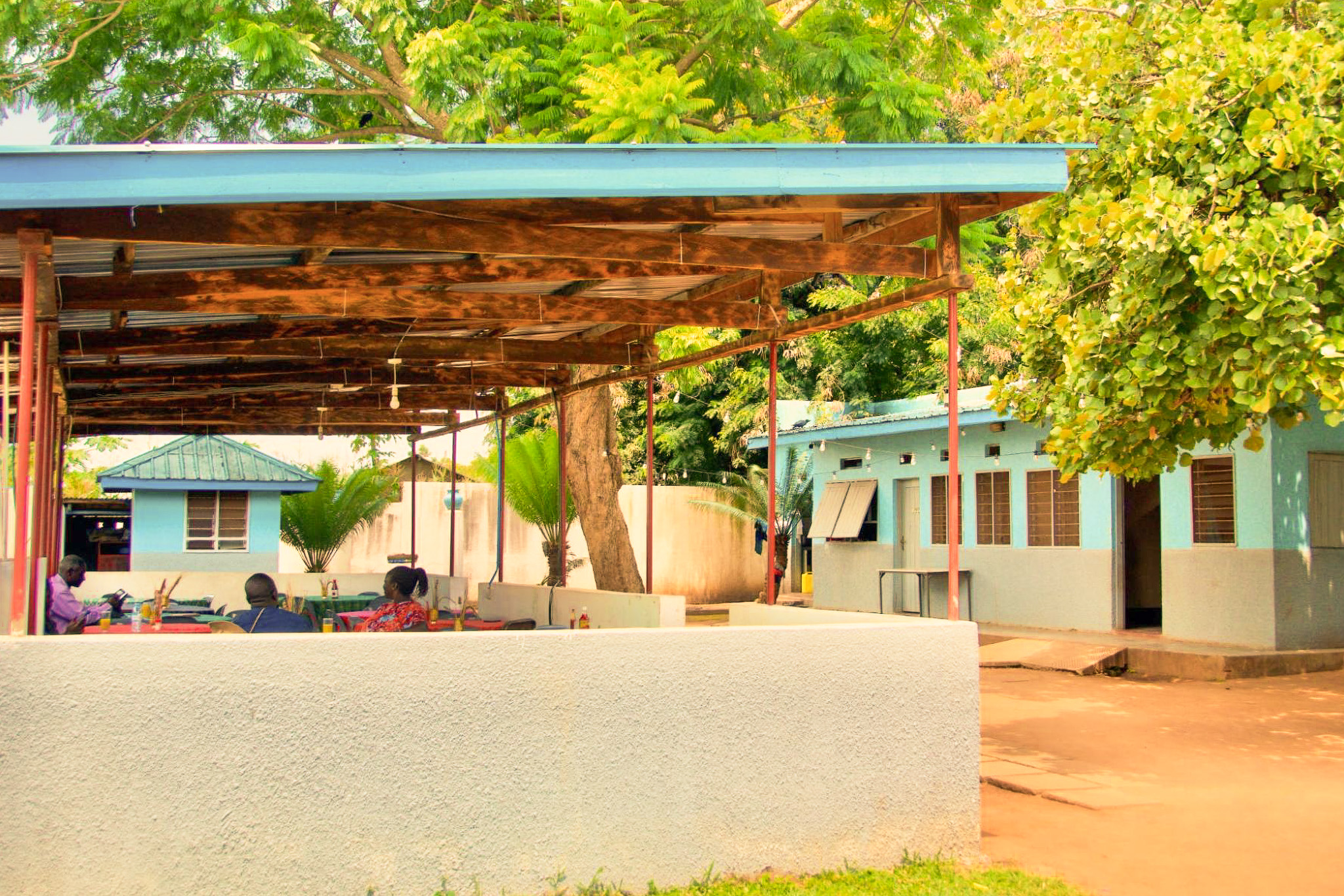 An outdoor seating area with a blue-roofed shelter and adjacent blue building, surrounded by lush green trees and people sitting at tables.