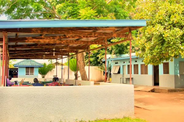 An outdoor seating area with a blue-roofed shelter and adjacent blue building, surrounded by lush green trees and people sitting at tables.