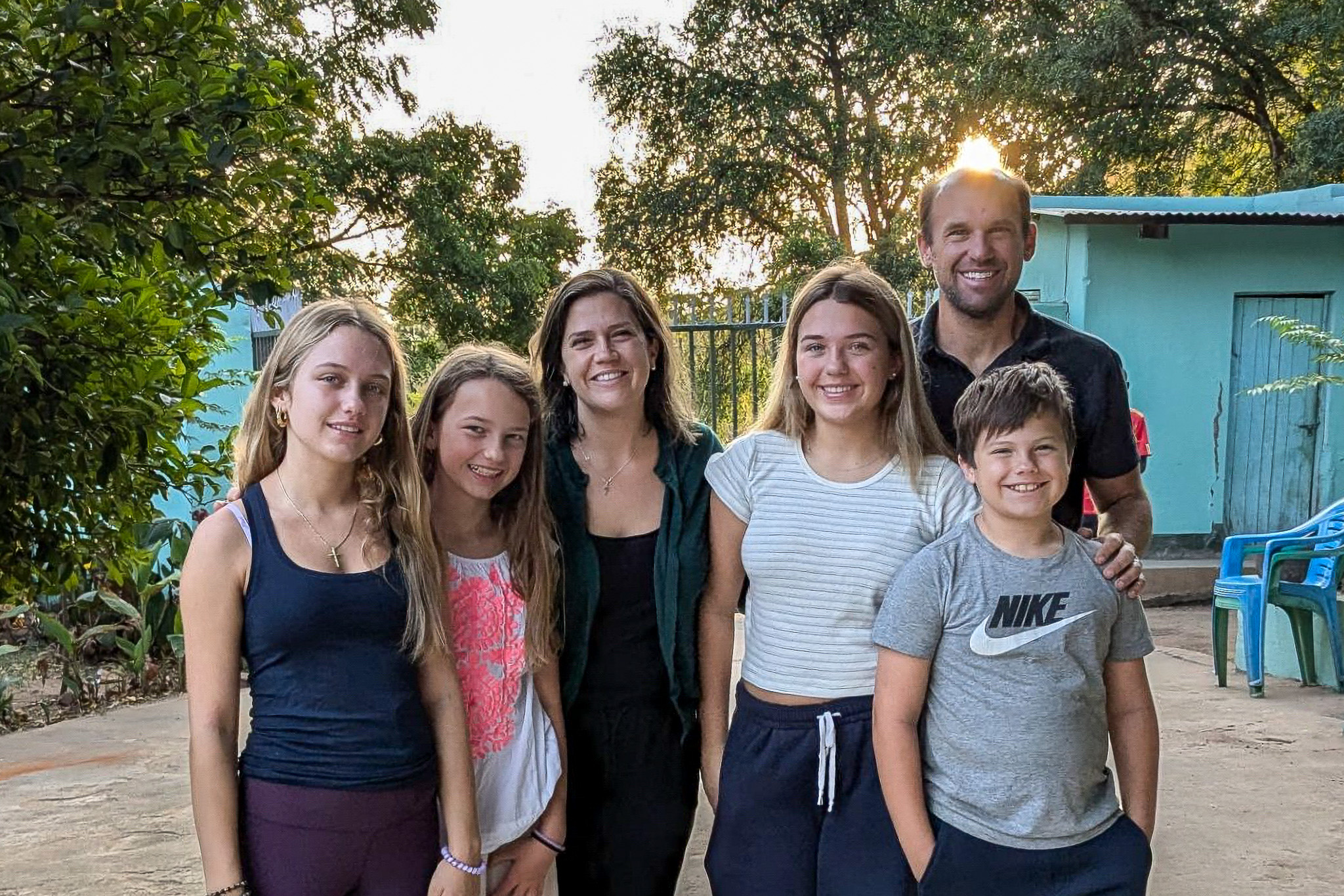 A group of six people, including four children and two adults, smiling outdoors with trees and a building in the background.