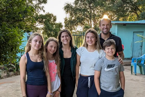 A group of six people, including four children and two adults, smiling outdoors with trees and a building in the background.