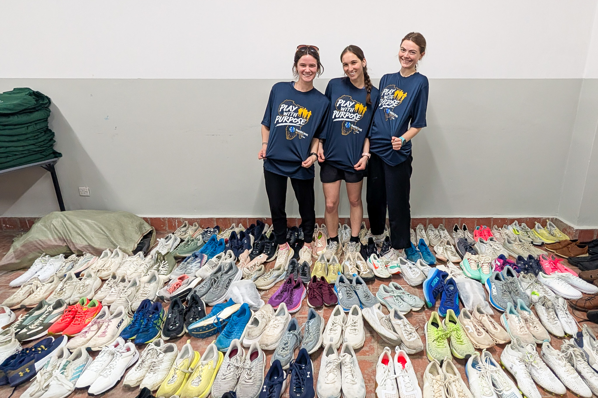 Three people in matching "Play with Purpose" shirts pose behind numerous pairs of neatly arranged sneakers on the floor.