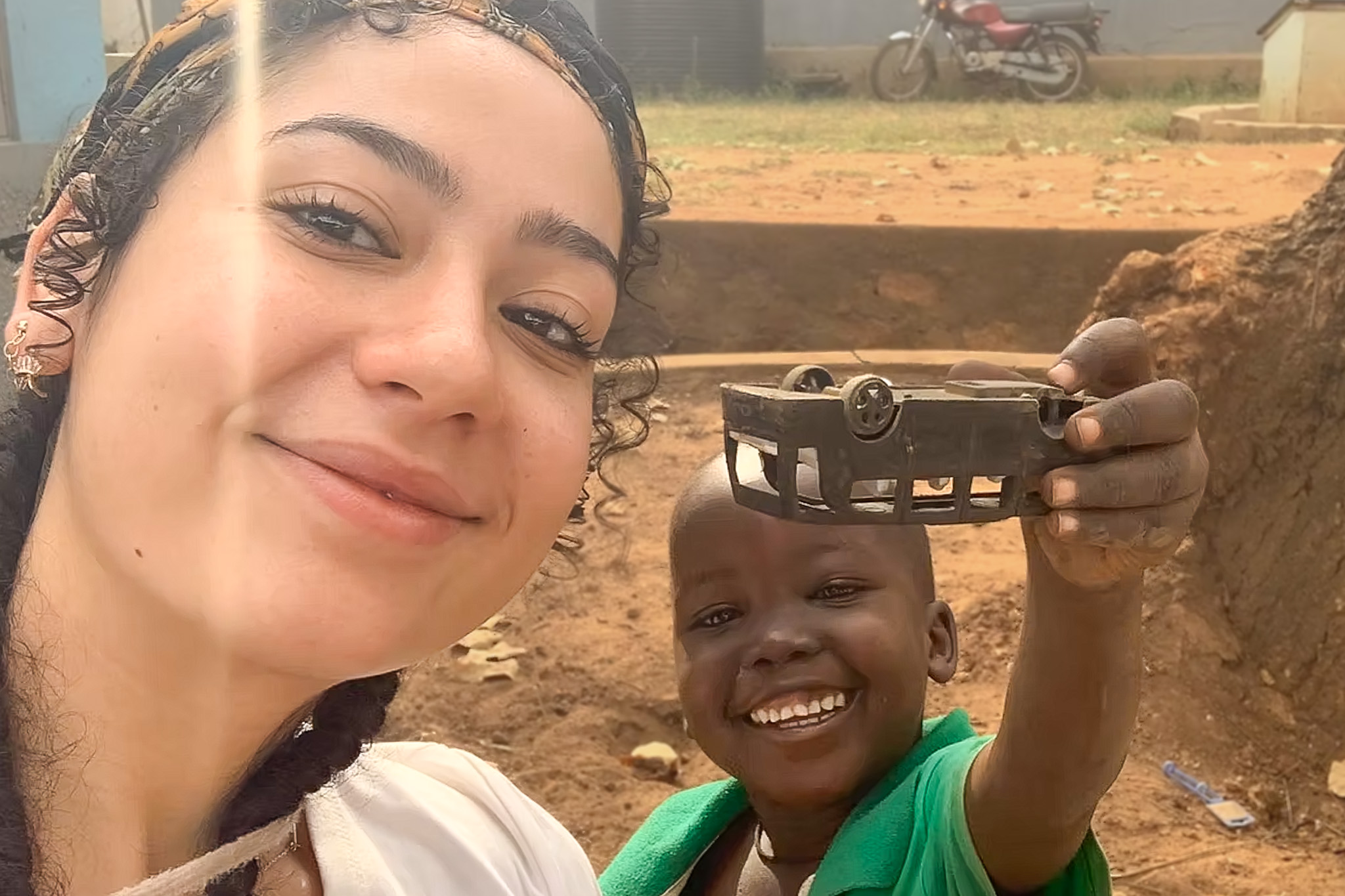 Smiling woman and child outdoors, child holding a toy car made from recycled materials, motorcycle and dirt road in the background.