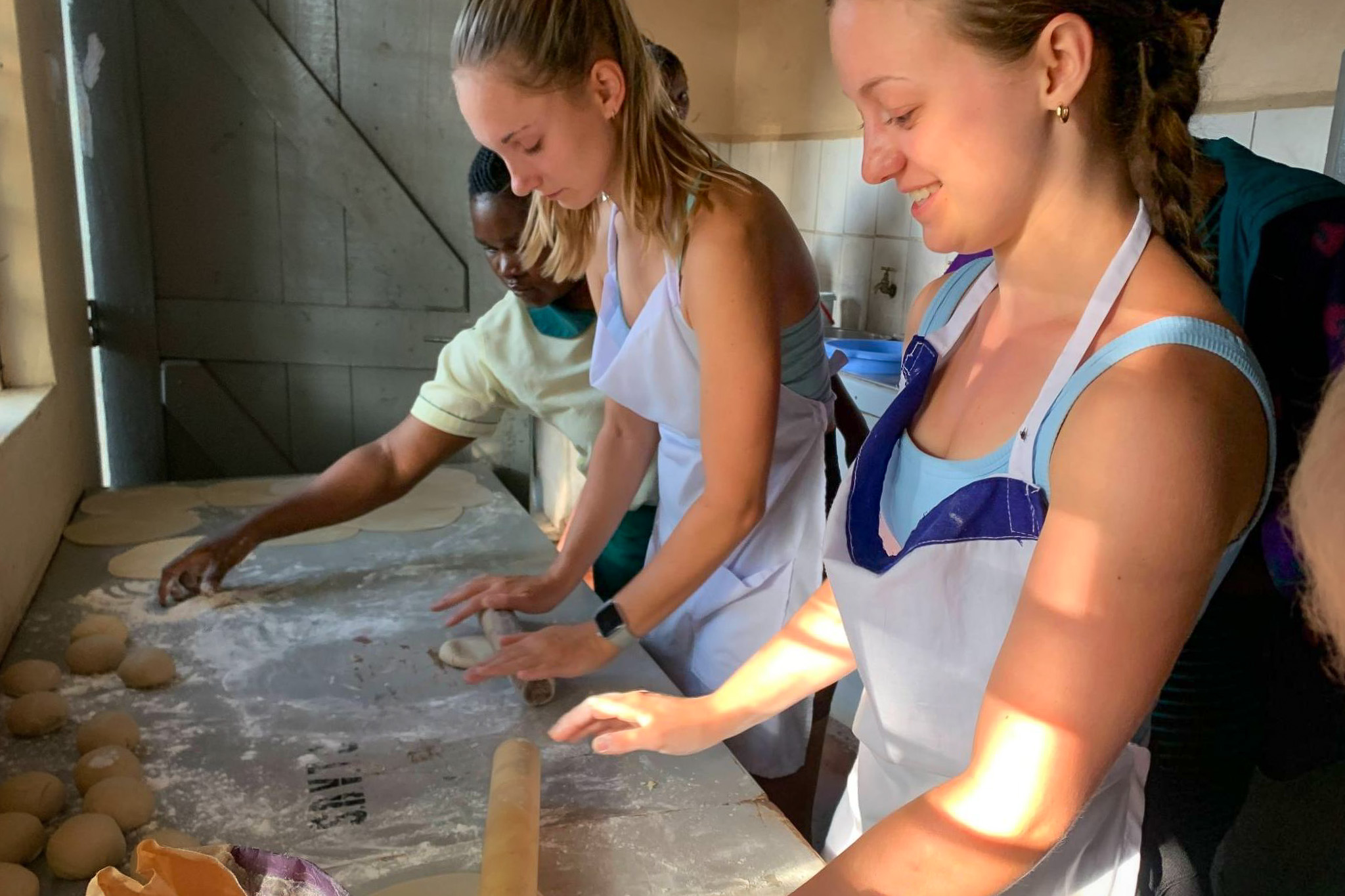 People in aprons rolling dough on a floured surface in a sunlit kitchen, engaging in a group baking activity.
