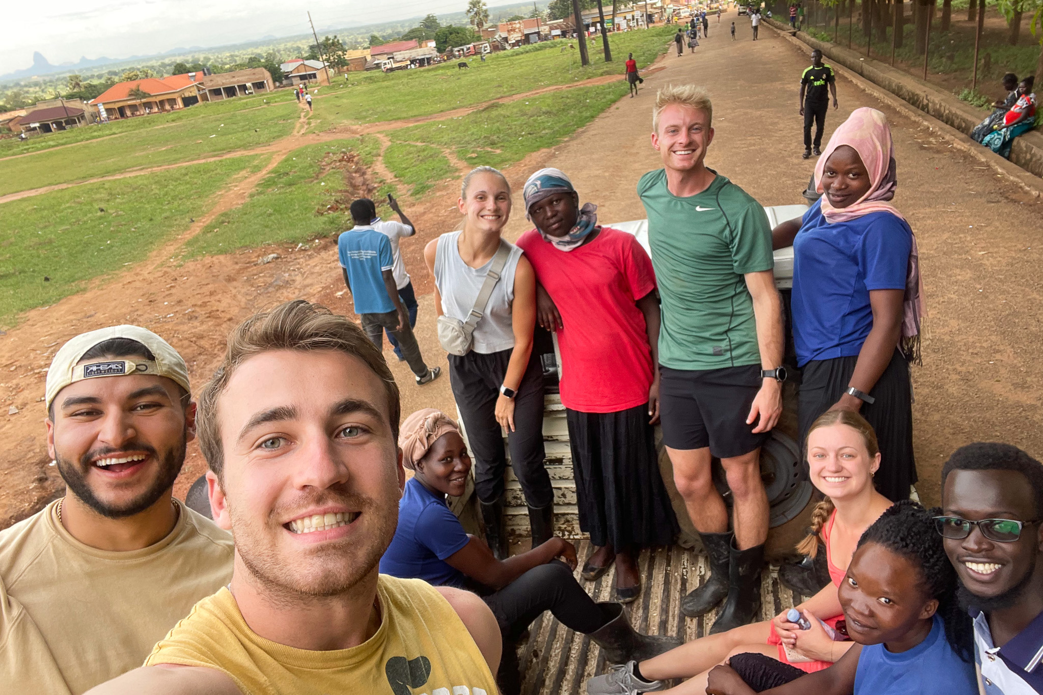 A group of cheerful people posing together outdoors on the bed of a truck, with a grassy field and buildings in the background.