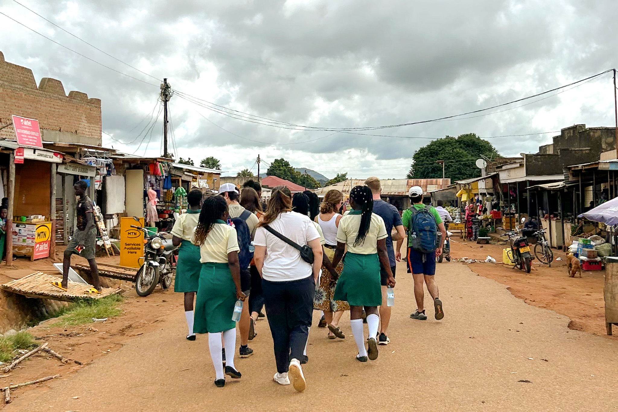A group of people, including schoolgirls in green uniforms, walk down a market street under a cloudy sky, surrounded by shops and motorcycles.