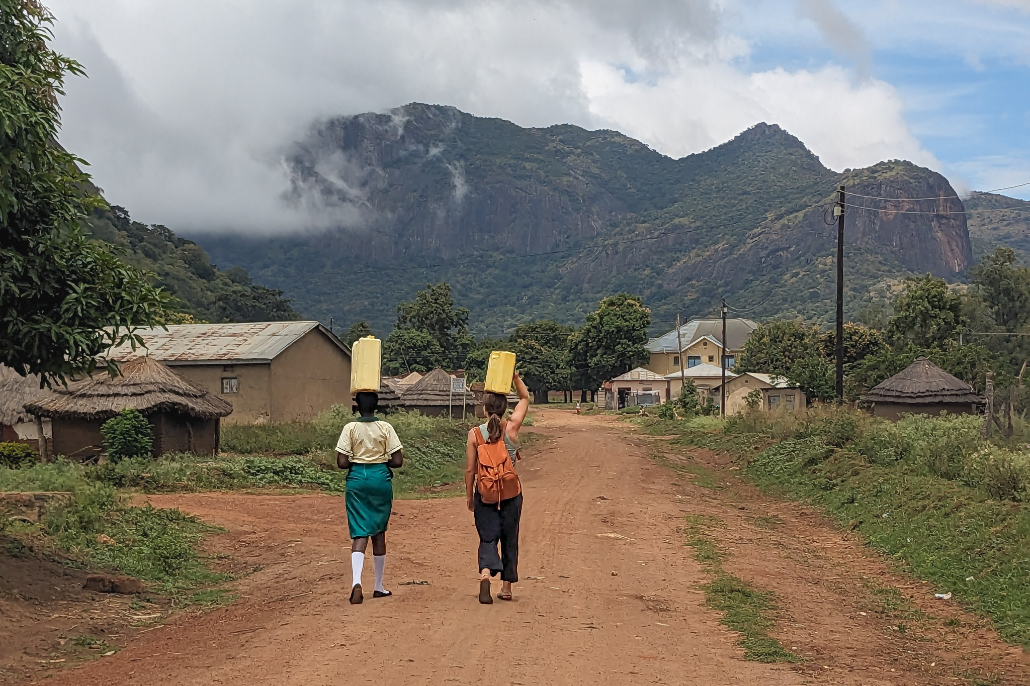 Two people walk on a dirt road carrying yellow jerry cans on their heads, with mountains and houses in the background.