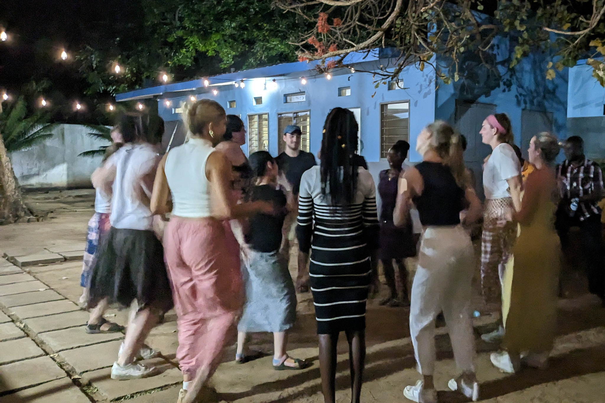 A group of people dancing together outside at night, under string lights, with a blue building in the background.